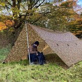 Person setting up the DD Hammocks 5x5 Tarp in a forest during autumn, showcasing its camouflage design.